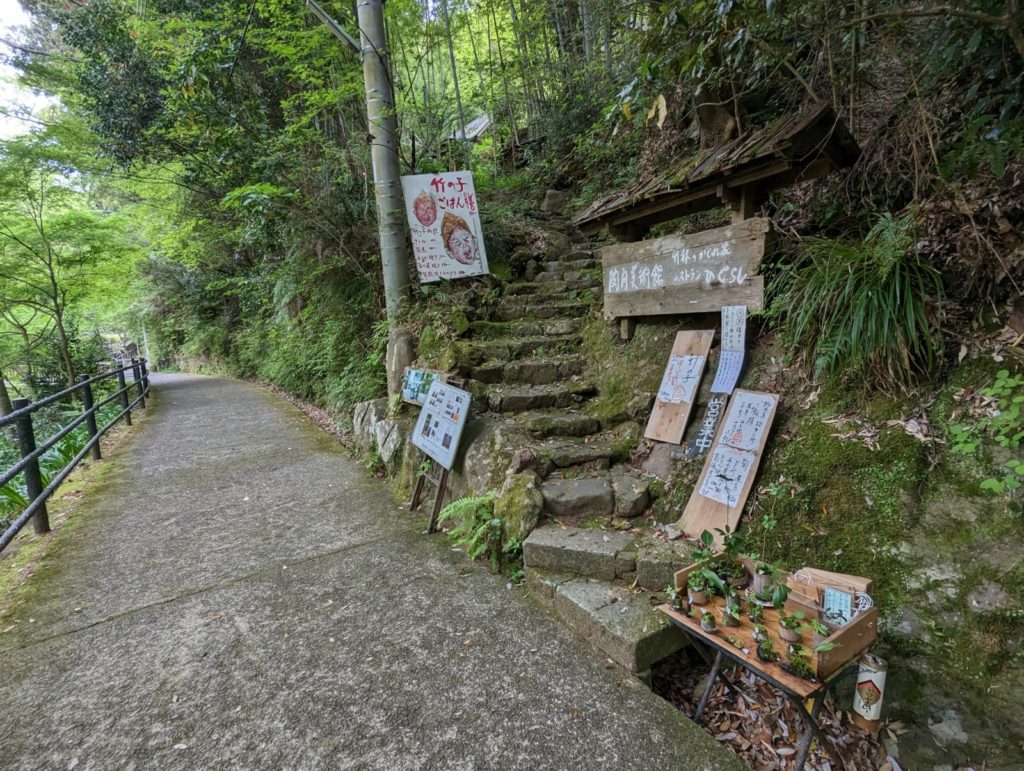 An einem Wanderweg führt eine Treppe hoch, an der allerlei Schilder auf ein Cafe aufmerksam machen.