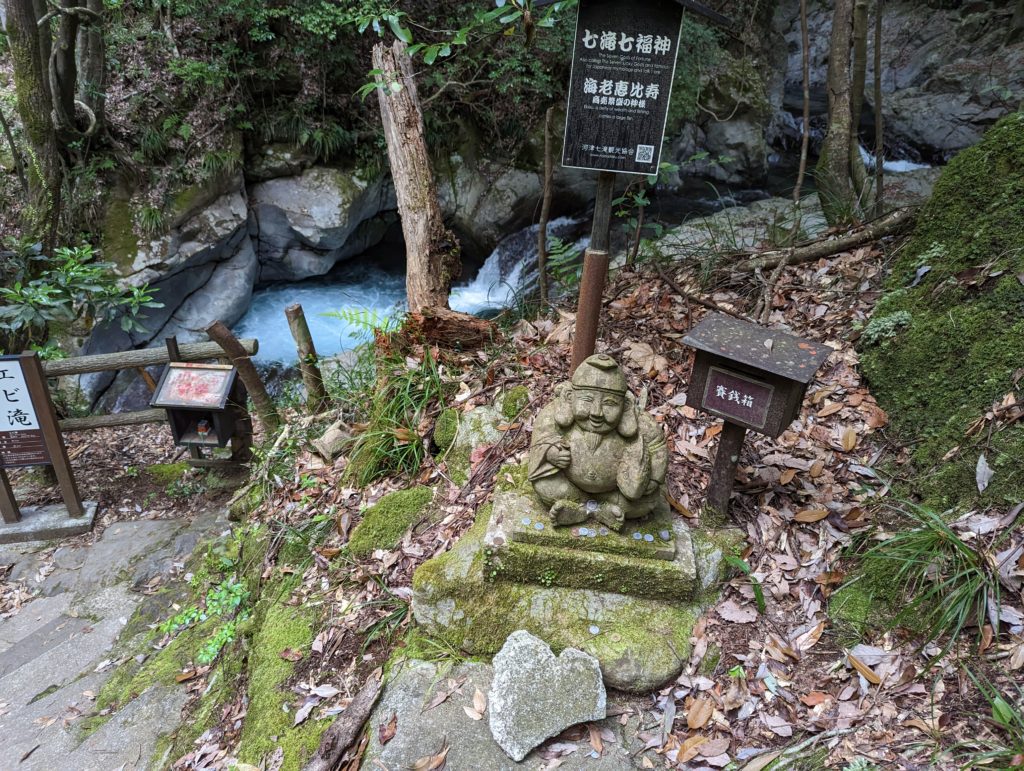 An einem Wasserfall ist eine kleine Statue die einen der sieben japanischen Götter des Glücks darstellt.