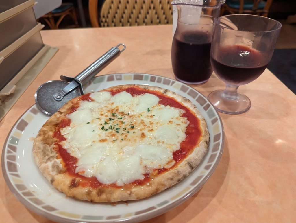 Eine Pizza mit viel Mozzarella und einem Pizzaschneider. Daneben ein gefülltes Weinglas und eine ebenfalls gefüllte Weinkaraffe.