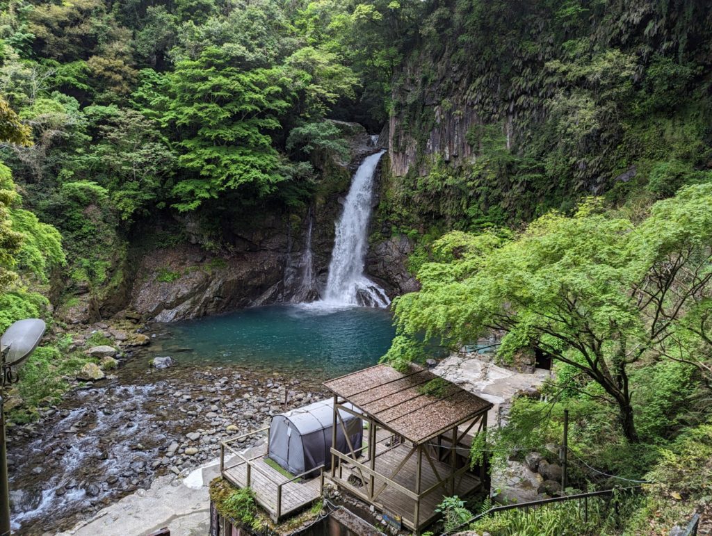 Ein großer Wasserfall ergießt sich in ein sonst sehr ruhiges GEwässer an dem auch ein Holzgebäude mit einem Zelt auf dem Dach steht.