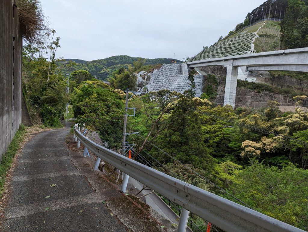 Von ener gemütlicheren Bergstraße sieht man eine große Brücke, die zu einem Tunnel führt. der Gebirgshang wurde mit Beton abgesichert.