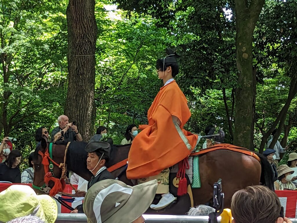 Historisch gekleidete Person auf einem Pferd reitet durch einen Park. Am Rand stehen etliche Zuschauer.