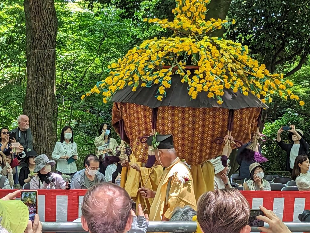 Eine Art Blumenschrein wird eine Straße entlang getragen. Am Rand stehen etliche Zuschauer.