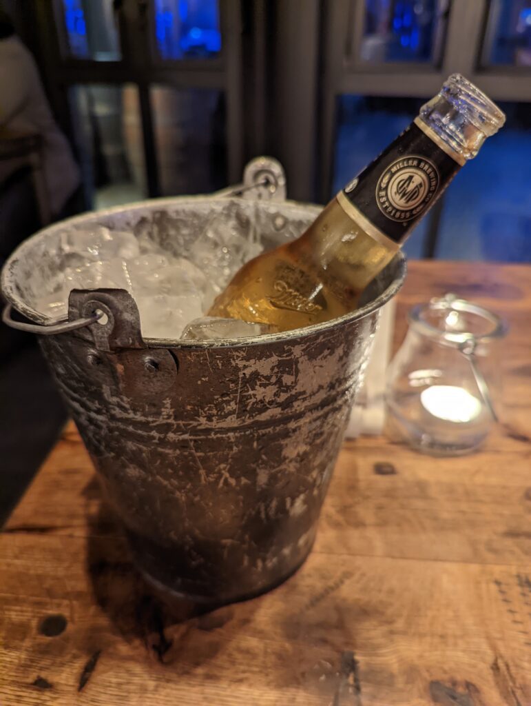 Eine Bierflasche serviert in einem kleinen rustikalen Eimer, der bis oben mit Eiswürfeln gefüllt ist.